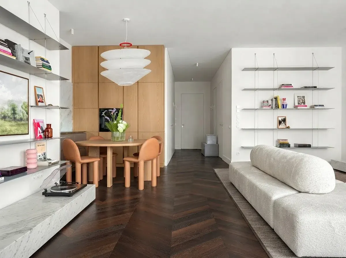 An open-plan apartment with a round oak dining table and orange chairs on the left, a white boucle sofa along the right wall, and a clear herringbone floor path running between them to the hallway door